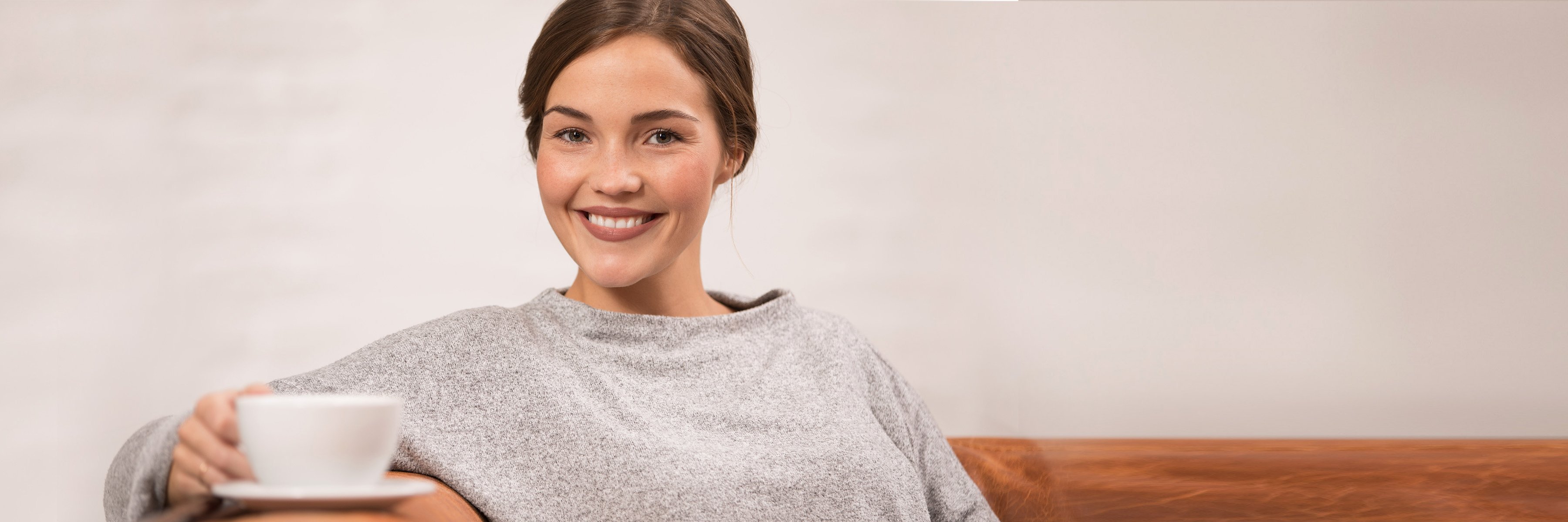 »Jestem zadowolona z mojego ubezpieczenia zdrowotnego.« Kobieta siedzi z uśmiechem na kanapie z filiżanką kawy.